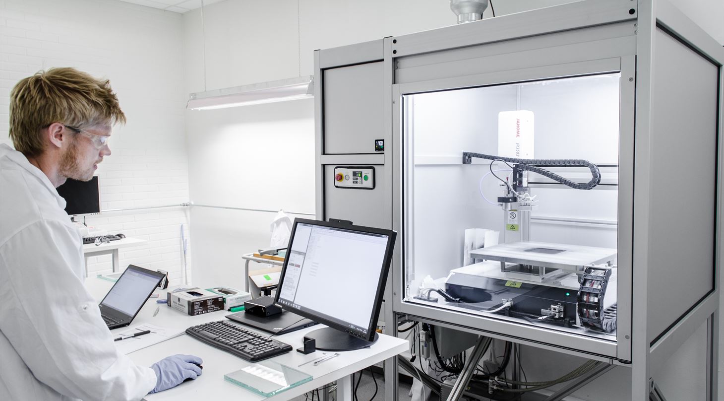 lab worker in front of a computer screen with a spray deposition robot in the background